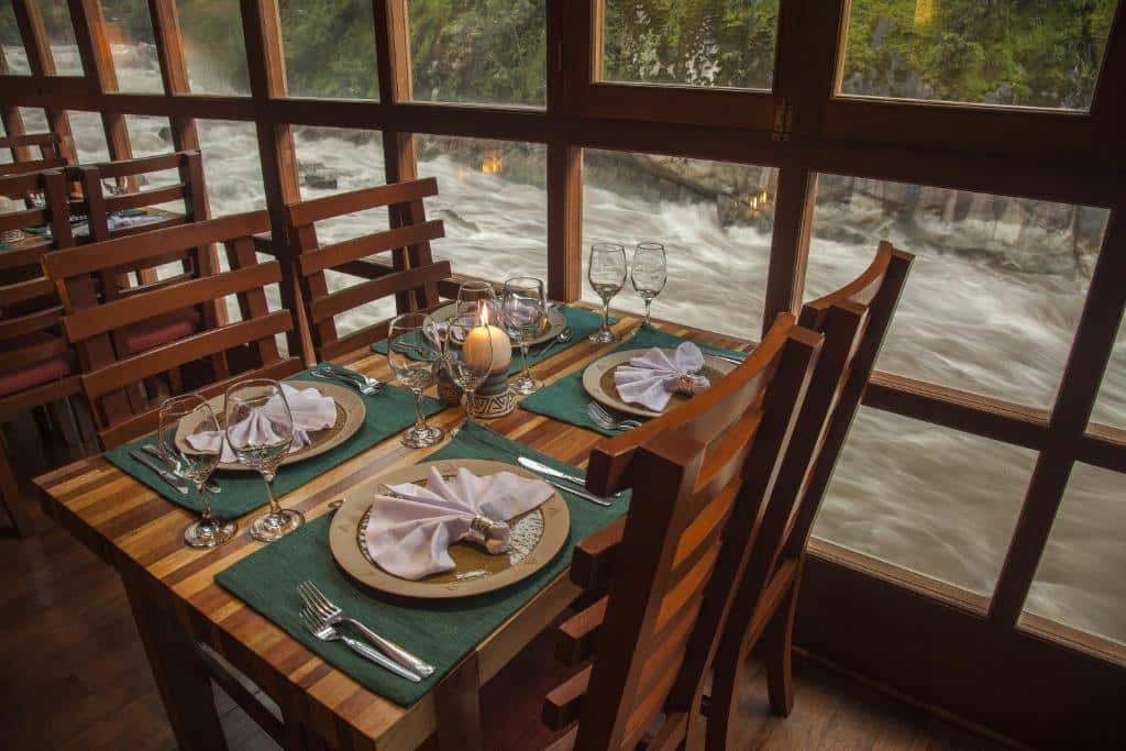 mesa para quatro pessoas com pratos e suplats e taças de vidro em cima, ao lado do rio Urubamba, visto pelas janelas amplas do restaurante.