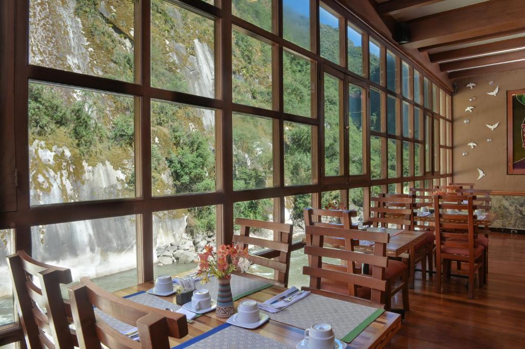 refeitório da casa del sol machupicchu com mesas de madeiras equipadas com xícaras e pratos brancos ao lado de amplas janelas que mostram a vegetação e o rio do lado de fora.