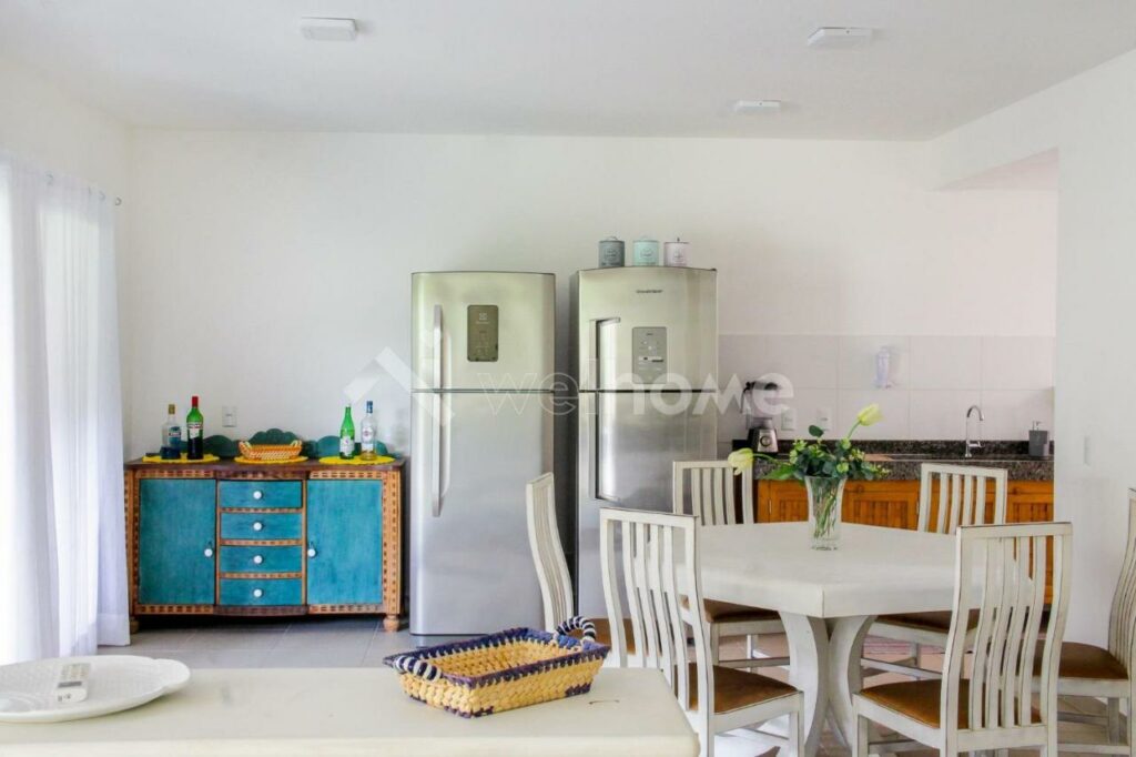 Cozinha da Casa em Condomínio na Praia em São Sebastião. Uma mesa quadrada com quatro cadeiras está em frente a uma bancada com pia e liquidificador. Ao lado da bancada há duas geladeiras, e um armarinho com bebidas está do outro lado das geladeiras.