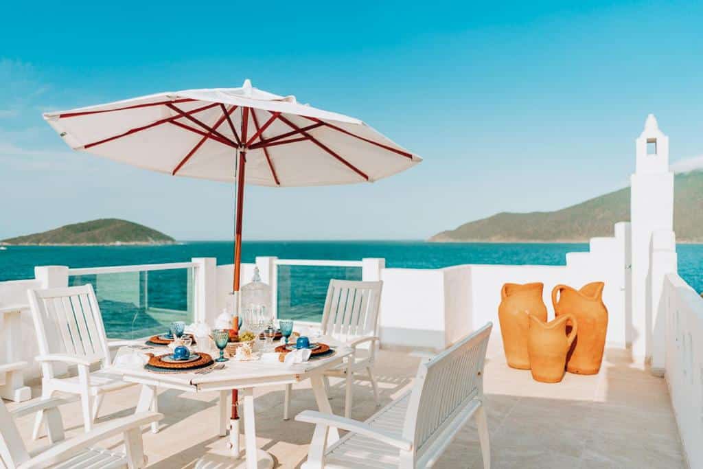 Área externa do airbnb Casa Grega. No canto esquerdo da imagem há uma mesa redonda com um guarda-sol preso ao meio e cadeiras em volta. Já no canto direito há três vasos decorativos de diferentes tamanhos. O parapeito da varanda é feito principalmente de concreto, mas no lado esquerdo há uma parte em vidro.