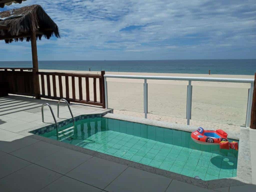 Área externa da Casa da Praia Pé na Areia. Uma piscina no meio com boias infantis, uma parede de vidro com vista para a areia da praia.