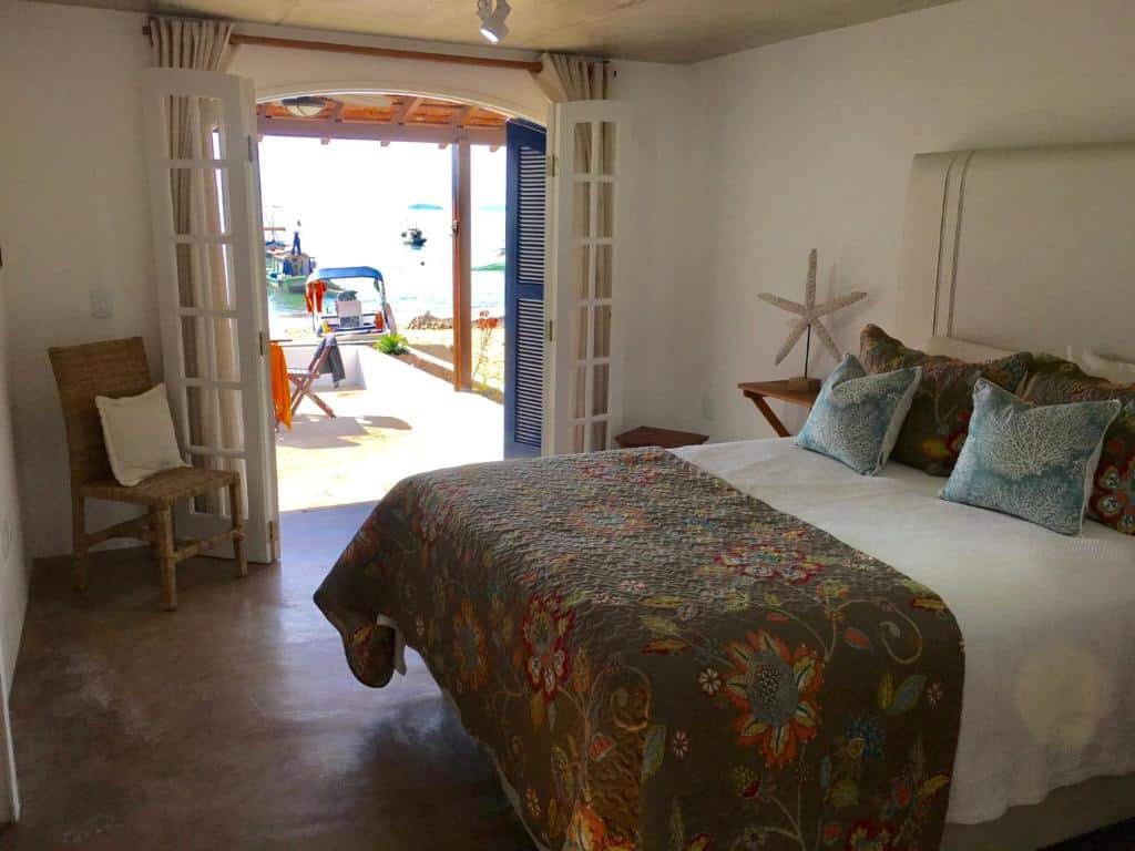Quarto do airbnb Casarao da Praia. No canto esquerdo do quarto há uma cadeira com uma almofada, no lado direito está a cama de casal e no fundo há uma grande porta que dá acesso ao quintal e a praia. Imagem para ilustrar o post airbnb em Ilha Grande.
