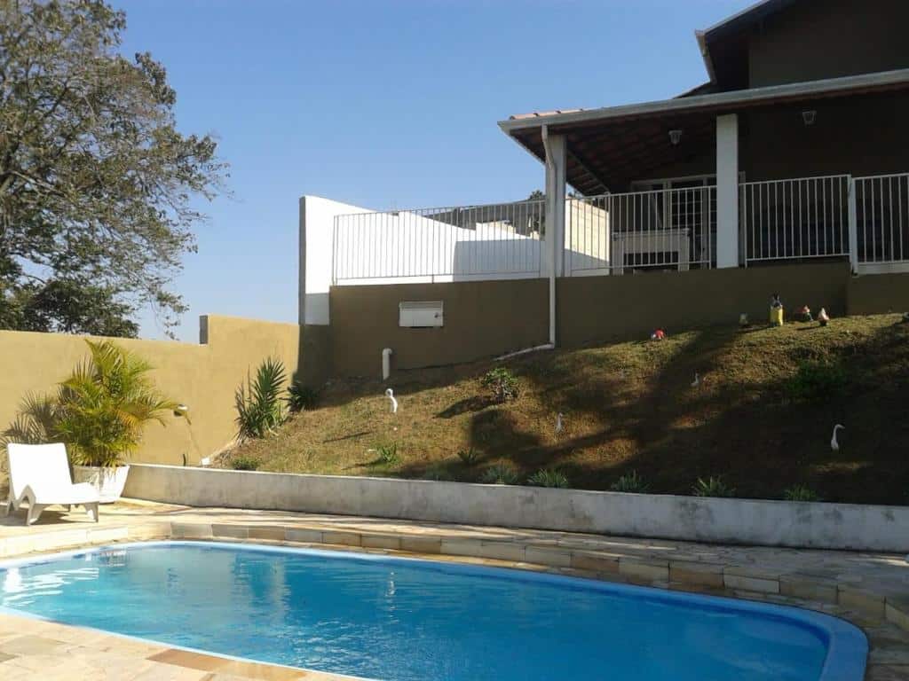 Uma piscina em um deck de pedra em Chácara Recanto Paraíso. Há uma espreguiçadeira no fundo à esquerda. Subindo um pequeno morro está a casa. 