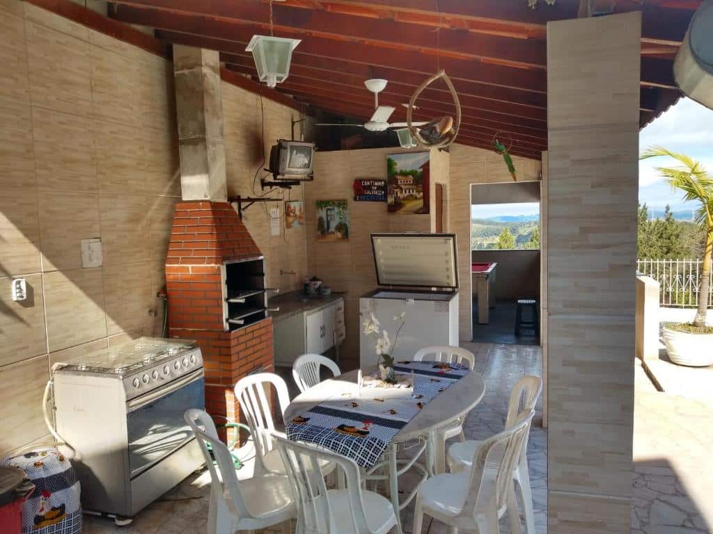 Foto da área de lazer em Chácara Recanto Paraíso. Há uma cozinha ao ar livre com mesa, cadeiras, fogão, churrasqueira, pia e freezer. Também há uma TV no topo de uma parede ao fundo. O rancho é coberto por um telhado.
Ilustra o post sobre airbnb em Serra Negra.