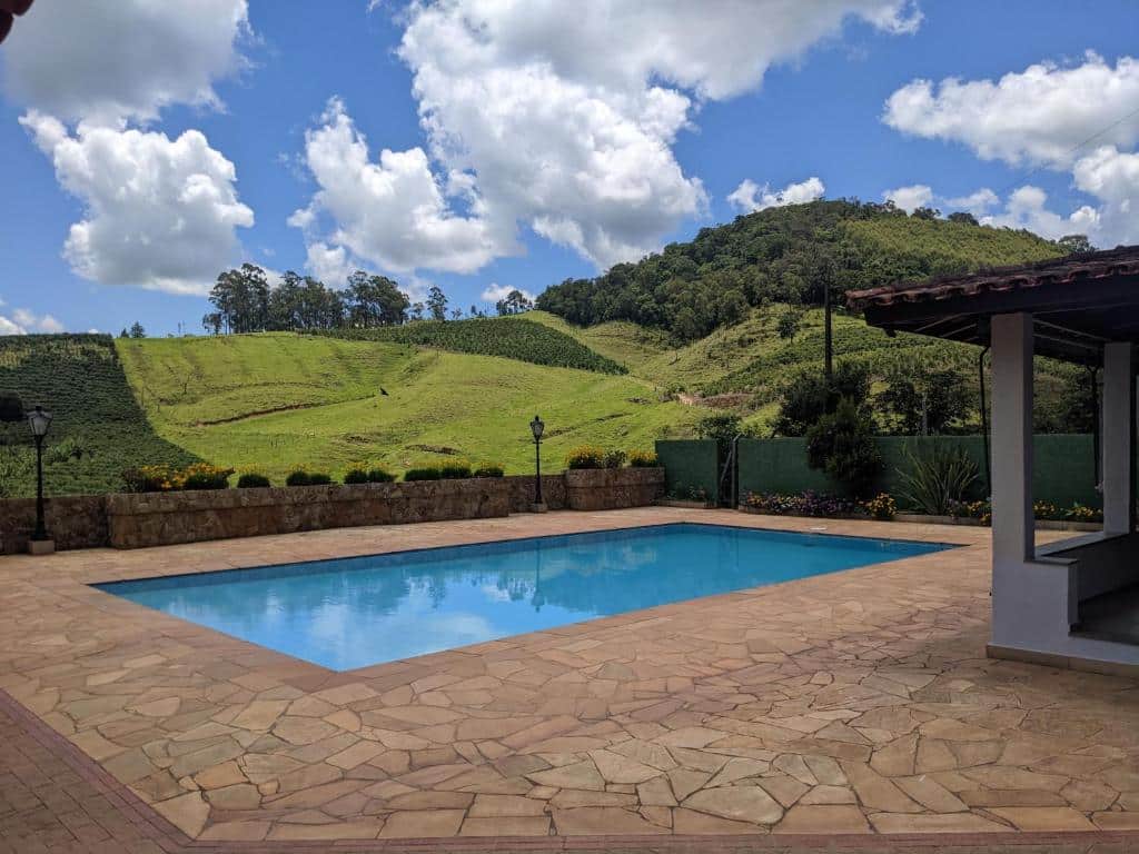 Grande deck com piscina em Chácara Santa Terezinha. O fundo mostra montanhas e árvores, e uma grama bem verde. O céu está azul com nuvens.