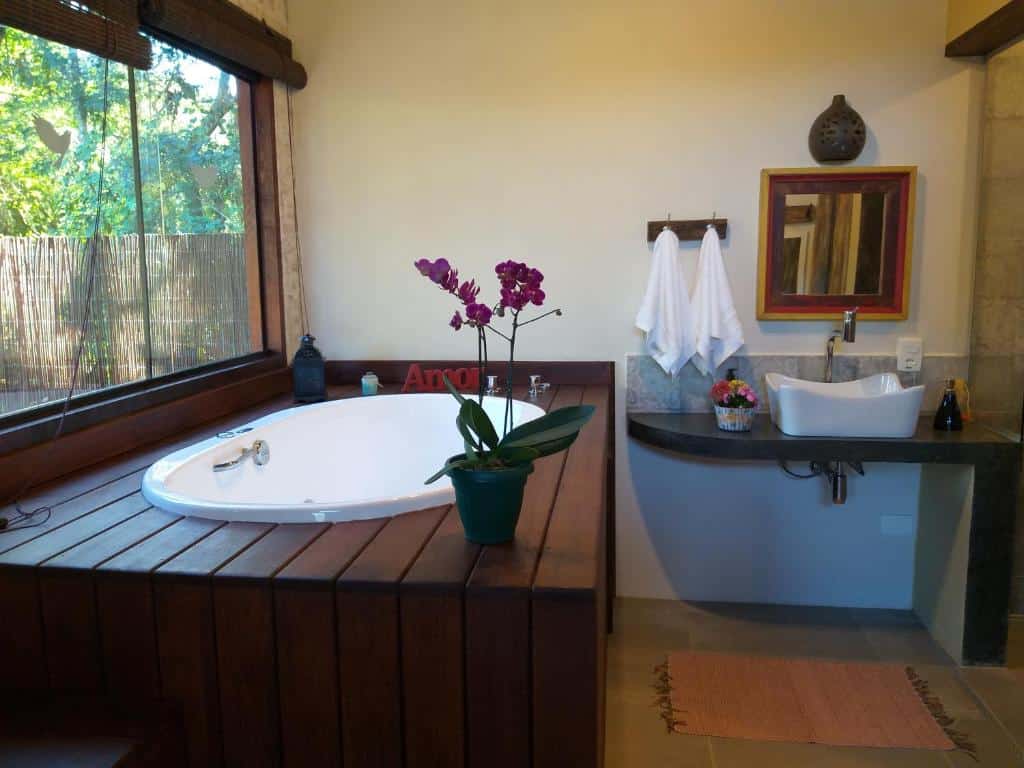 Banheiro do Chalé Alecrim. Uma banheira de hidromassagem está do lado esquerdo, ao lado de uma janela. Do lado direito está uma pia e um espelho quadrado na parede.