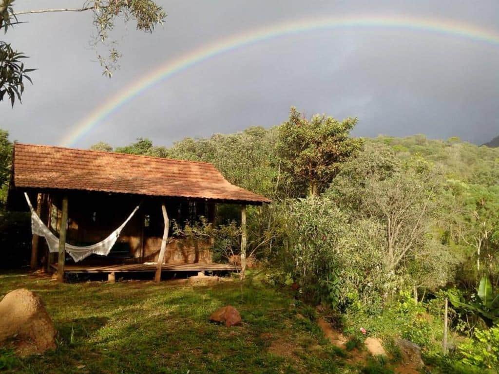 Fachada do Chalé Candeia no Matutu que mostra do lado esquerdo uma casinha rústica de madeira com uma rede e do lado direito várias árvores verdes. Imagem ilustrando post Airbnb em Aiuruoca.