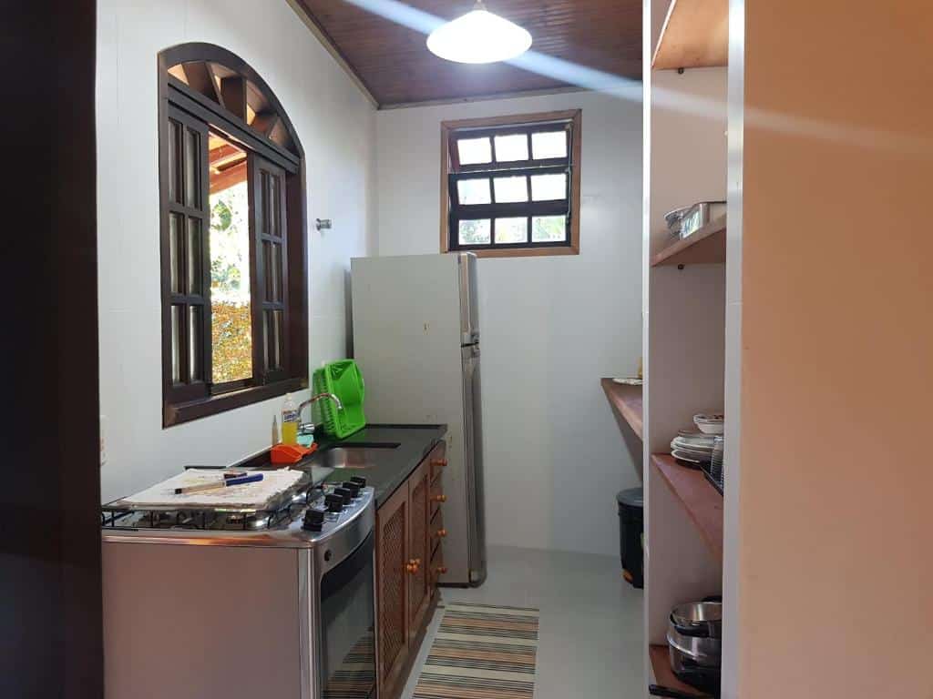 Cozinha de um dos Chalés e flats Recanto Primavera. Encostados na parede esquerda estão um fogão, um armário com pia e uma geladeira. Há uma janela logo acima da pia. No lado direito estão as bancadas e um armário com utensílios.