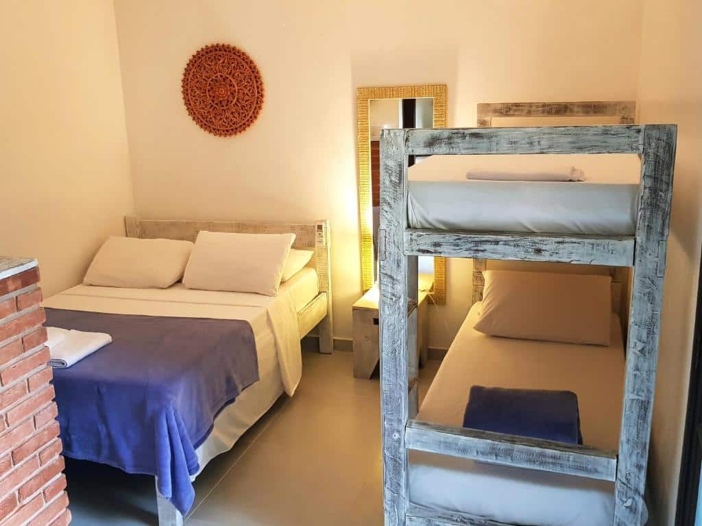 Quarto de um dos Chalés e flats Recanto Primavera, um dos airbnb em Camburi. Ao lado esquerdo está uma cama de casal separada da beliche do lado direito por uma mesinha com abajur e um espelho. Todas as camas tem travesseiros, lençol e cobertor.