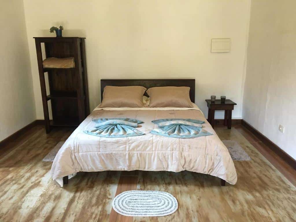 Foto do quarto do Chalé Filhos da Terra. Representa o post sobre airbnb em São Francisco Xavier. cama de casal está no centro da imagem. Ao lado esquerdo dela há uma estante de madeira, e ao direito, um pequena mesa de cabeceira. É espaçoso.