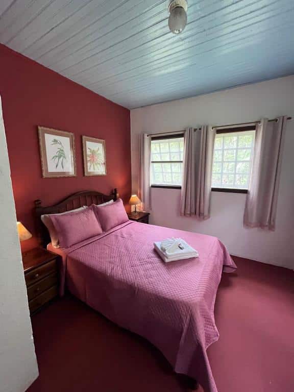 Quarto do airbnb Chalé Gaia. A cama de casal está centralizada no quarto e encostada na parede do lado esquerdo. Em cada lado da cama há uma mesa de cabeceira com um abajur em cima. Na parede do lado direito há duas janelas com cortinas. Imagem para ilustrar o post airbnb em Itatiaia.