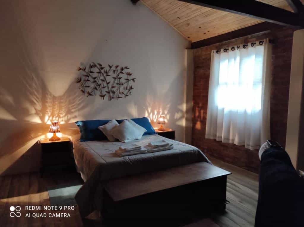 Foto do quarto do Chalé Lavanda com Hidro. Representa o post sobre airbnb em São Francisco Xavier. A cama box de casal está ao centro. Dos dois lados dela há uma mesa com cabeceira e abajur. Na direita há uma janela com cortina.