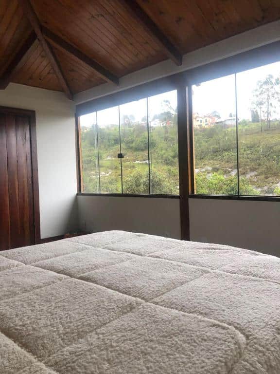 Foto do quarto do Chalé Lírio do Campo. Ilustra o post sobre airbnb em Ouro Preto. O ângulo que vemos é como o de quem está deitado na cama. À nossa frente há duas janelas de vidro com vista para a vegetação. À esquerda há uma porta.