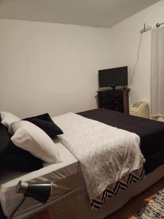 Quarto do airbnb Chalé no centro de Penedo. No lado esquerdo está encostada a cama de casal e no lado direito há uma pequena cômoda com uma TV em cima, um ar-condicionado de chão e é possível ver parte de uma cortina também. Imagem para ilustrar o post airbnb em Itatiaia.