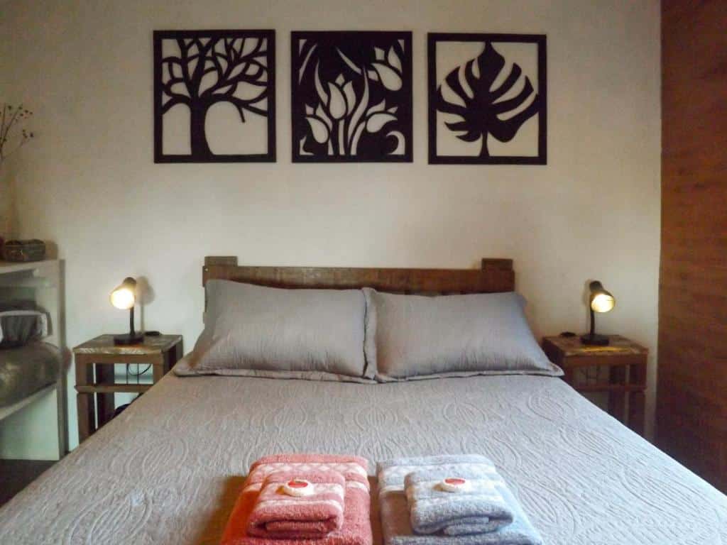 Foto do quarto do Chalé rústico na natureza de SFX. Representa o post sobre airbnb em São Francisco Xavier. A cama de casal está no centro da foto. Dos dois lados dela há uma mesa de cabeceira com abajur.