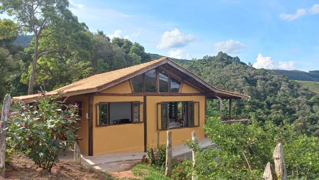 Parte externa do Chalé Sabiá que mostra no meio da foto a construção da casa com paredes amarelas, duas janelas abertas, detalhes em vidro e madeira e telhado pontudo. Em volta da casa tem bastante árvores verdes e uma vista para as montanhas durante o dia. Imagem ilustrando post Airbnb em Aiuruoca.