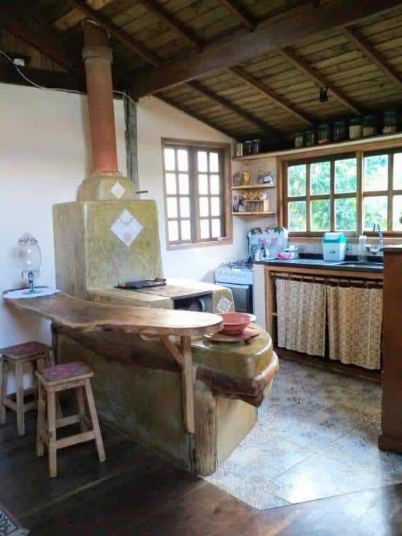 Cozinha do Chalé Solar no Matutu com uma bancada de madeira e duas banquetas, ao lado um fogão a lenha e no fundo perto das janelas tem um fogão a gás com prateleiras em cima na parede e do lado do fogão tem a pia de levar a louça. Imagem ilustrando post Airbnb em Aiuruoca.