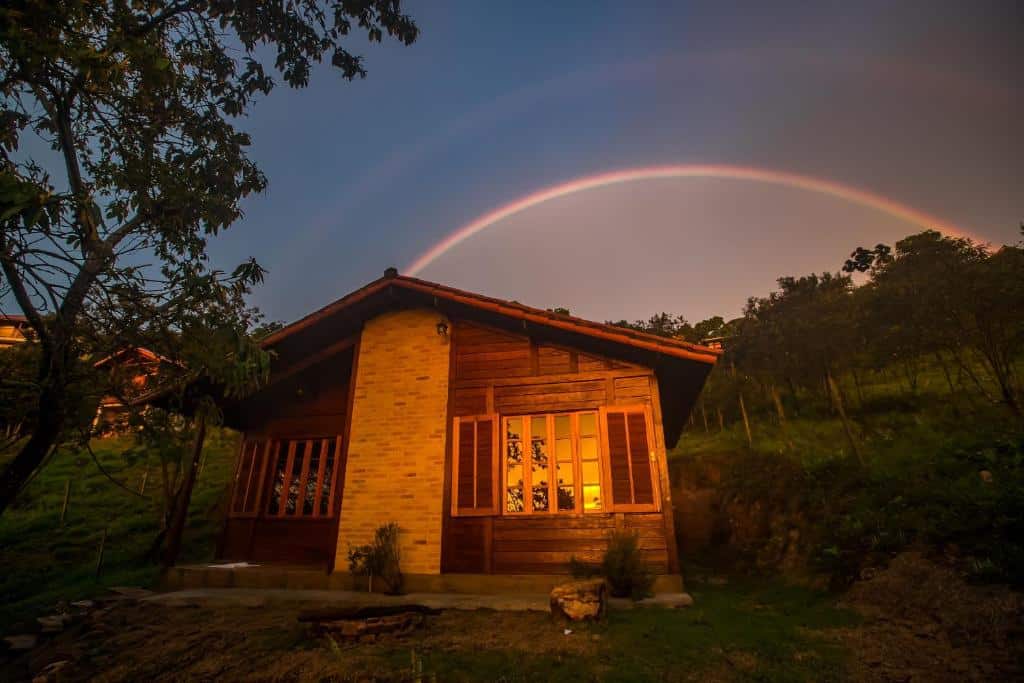 Fachada de um dos Chalés Belo Vale - Ibitipoca que mostra uma casa com duas janelas, paredes de madeira e tijolos. Em volta há bastante gramado e árvores e o céu está azul com um arco-íris. Imagem ilustrando post Airbnb em Ibitipoca.