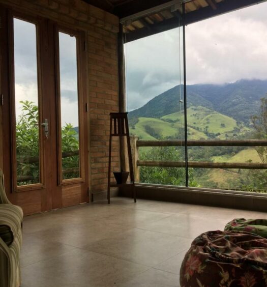 Imagem tirada de dentro da sala do Chalés de Valfena em SFX. A vista é para montanhas. Há um sofá no canto esquerdo e puffs na direita.