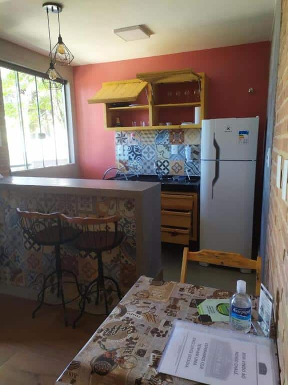 Parte do Chalés Estrada Real que mostra uma mesa de dois lugares de madeira e ao fundo uma bancada de concreto com azulejos coloridos e duas banquetas separando a sala da cozinha. Na cozinha tem uma geladeira na parte direita, uma pia com um armário em baixo e um armário em cima com copos dentro, e do lado esquerdo tem uma janela de vidro. Imagem ilustrando post Airbnb em Ibitipoca.