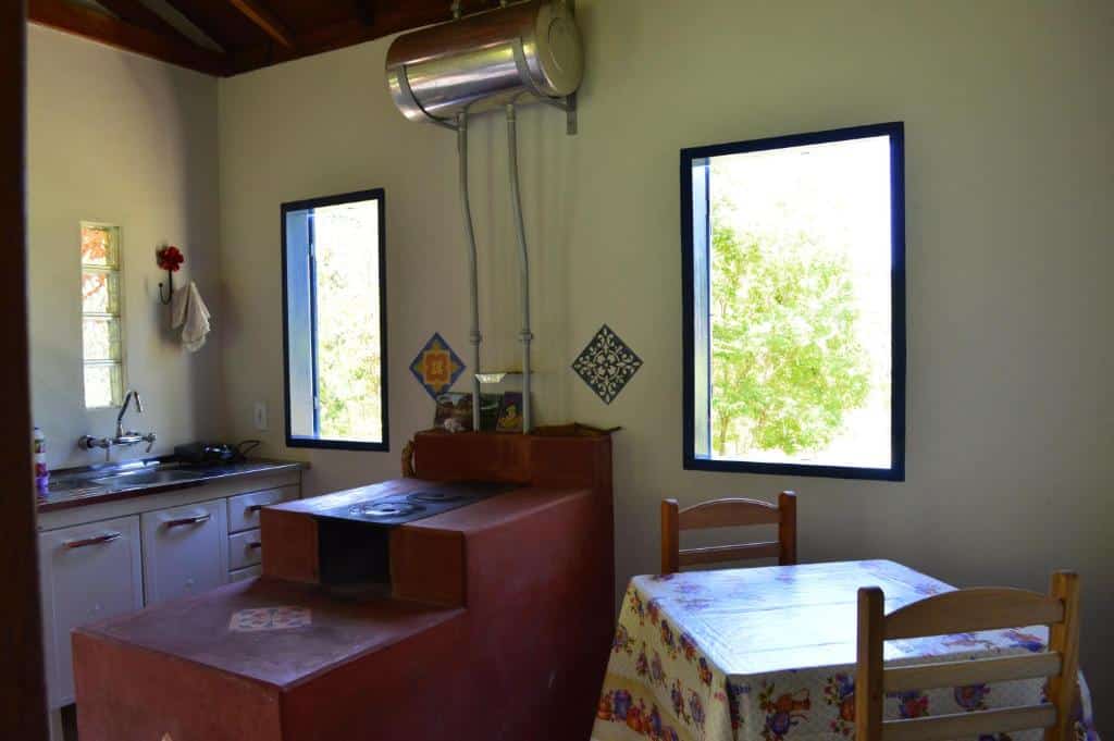 Cozinha do Chalés Serrinha do Papagaio com uma mesa de dois lugares e uma janela na parte direita, Já do lado esquerdo tem um fogão a lenha e ao fundo uma pia para lavar louças com uma janela aberta ao lado. Imagem ilustrando post Airbnb em Aiuruoca.