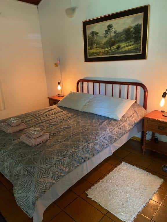 Foto do quarto do Chalés SFX. Representa o post sobre airbnb em São Francisco Xavier. A cama é de casal e está no centro da foto. Dos dois lados dela há uma mesa de cabeceira com abajur.