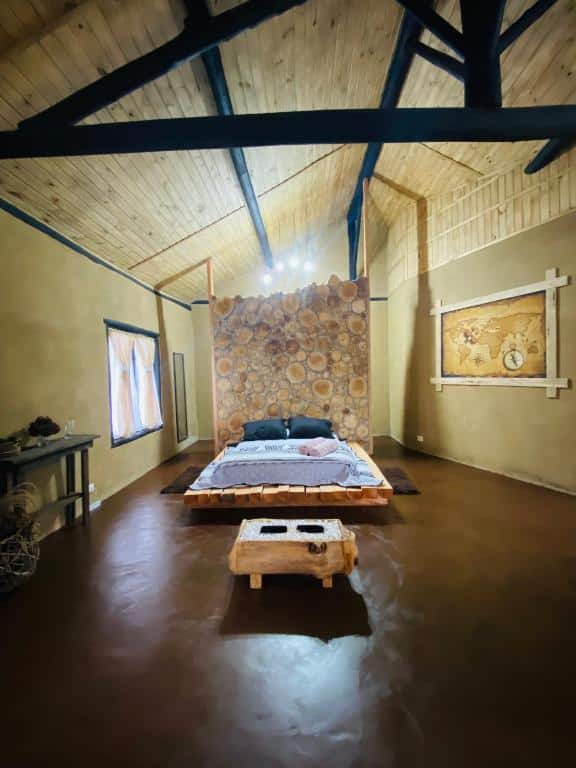 Foto do quarto do Chalé SPAço Altos da Serra. Representa o post sobre airbnb em São Francisco Xavier. A cama está no centro, sobre uns pallets de madeira. O quarto é bem espaçoso, o teto é alto. À esquerda da cama há uma janela, e atrás dela há uma divisória.