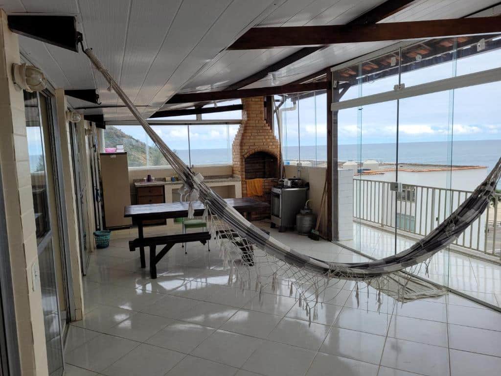 Varanda da Cobertura vista mar. Uma rede na frente, atrás uma mesa de madeira, com uma churrasqueira e uma cozinha completa. Uma porta de vidro do lado direito com acesso a sacada e vista para o mar.