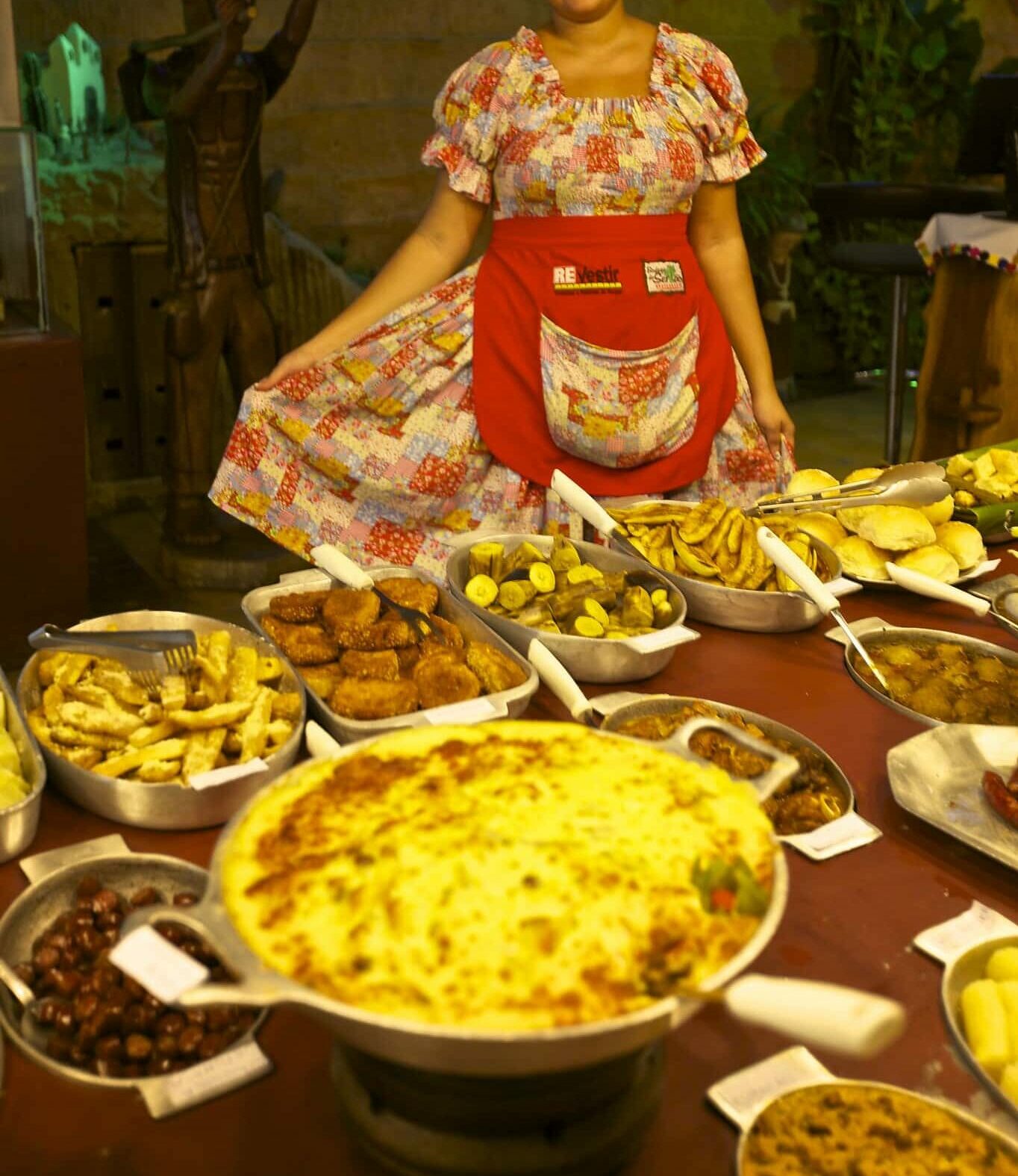 mulher com uma roupa típica de maceió, segurando a saia, atrás de uma mesa cheia de pratos tipicos. Representa restaurantes em Maceió