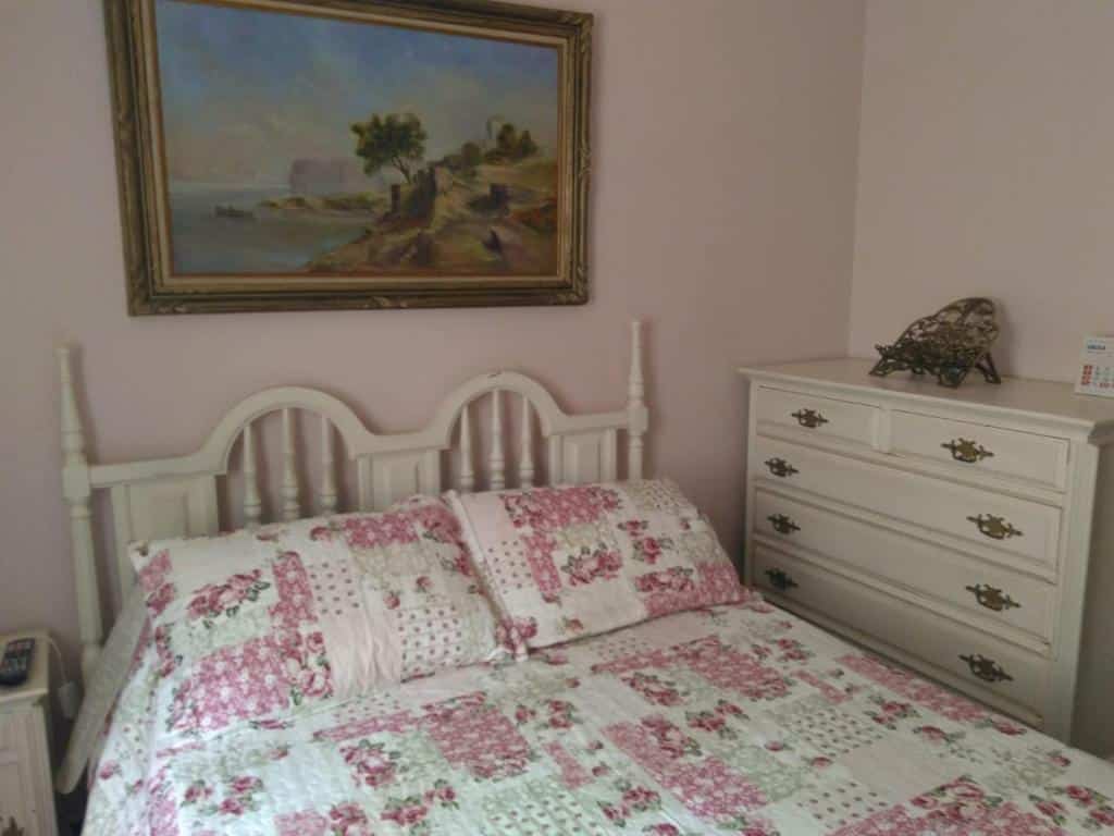 Foto do quarto em Condomínio Vila Paradiso. As paredes são rosa claro. Há uma cama no centro com cabeceira de madeira branca, e ao seu lado na direita há um gaveteiro branco de madeira. Há um quadro atrás da cama, decorando a parede.
Ilustra o post sobre airbnb em Serra Negra.