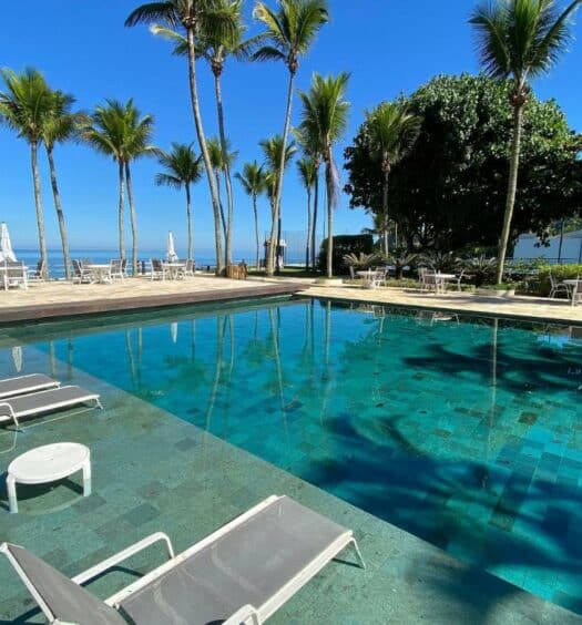 Ampla piscina cercada por espreguiçadeiras e palmeiras no Condomínio pé na areia, um dos airbnb em Barra do Una. Ao fundo é possível ver o mar.