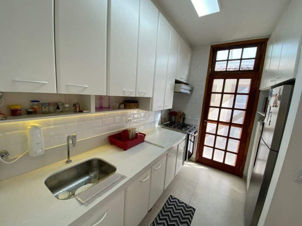 Cozinha do Condomínio pé na areia repleta de armários e bancadas brancas, onde fica uma pia. Ao fundo está um fogão que encara uma geladeira. Há uma porta no final do corredor entre os móveis.