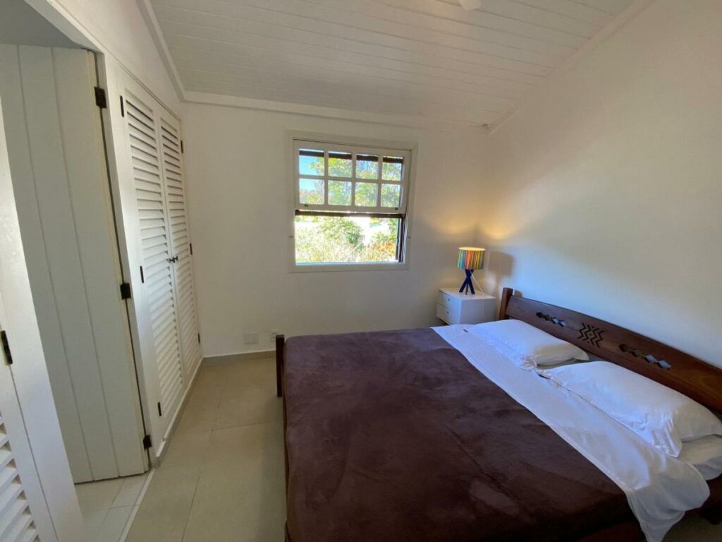 Quarto do Condomínio pé na areia, um dos airbnb em Barra do Una. Uma cama de casal com mesinhas de cabeceira e abajur dos dois lados está encostada na parede direita e encara um armário na outra parede. Ao lado esquerdo da cama há uma janela aberta.