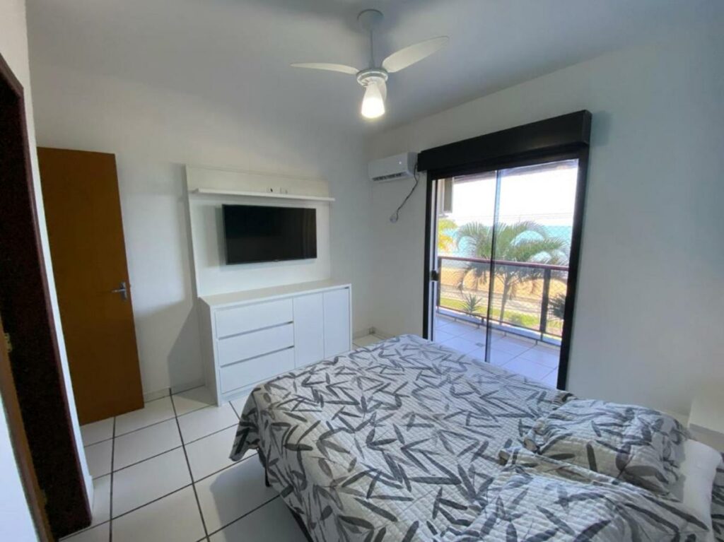 Quarto do Condomínio San Martim, um dos airbnb em Caraguatatuba. Uma cama de casal com colcha e travesseiros está encostada na parede ao lado direito e encara uma cômoda com TV na parede oposta. Na parede ao lado da cômoda há uma porta de vidro que leva à varanda com vista para a praia. Há um aparelho de ar-condicionado ao lado da porta.
