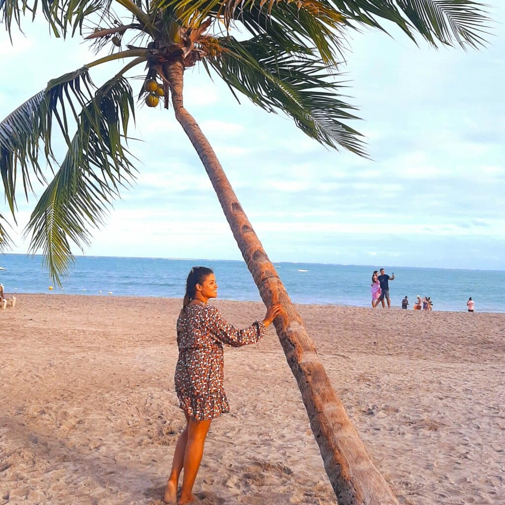 Mulher de costas, olhando para o lado e sorrindo, com uma das mãos encostada em um coqueiro inclinado na praia de Antunes