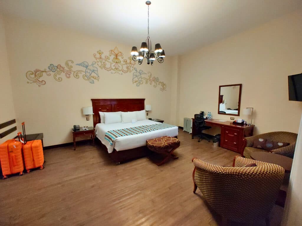 suíte do Costa del Sol Wyndham Cusco com uma grande cama de casal no centro do quarto, um candelabro pendurado no teto sobre a cama, uma mesa de madeira com cadeira ergonômica à direita da imagem e um conjunto de malas laranjas à esquerda.