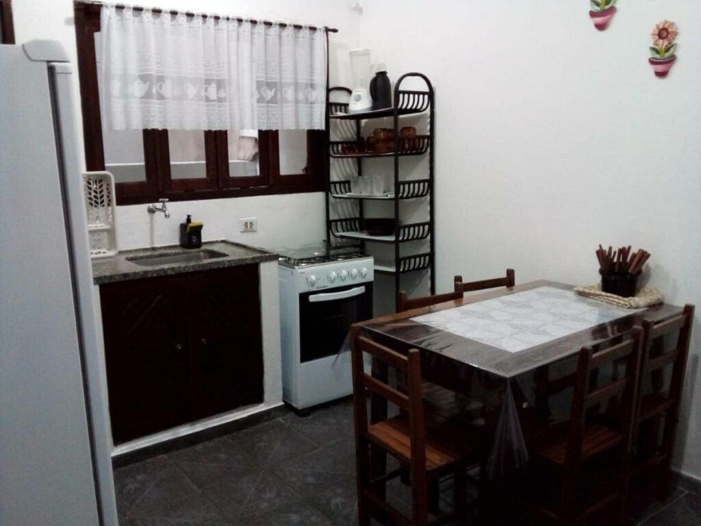 Cozinha do Excellence Apart com mesa de jantar com cinco cadeiras do lado direito, ao fundo um fogão e uma pia.