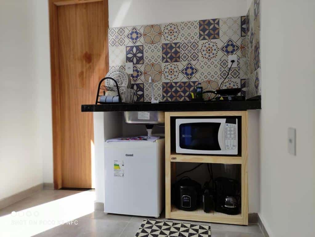 Cozinha do airbnb Flat Container Pontal do Atalaia. No lado direito da imagem há uma pia com um frigobar em baixo e uma prateleira com um micro-ondas, uma cafeteira, uma sanduicheira e uma panela elétrica também em baixo da pia. No lado esquerdo há uma porta de madeira.