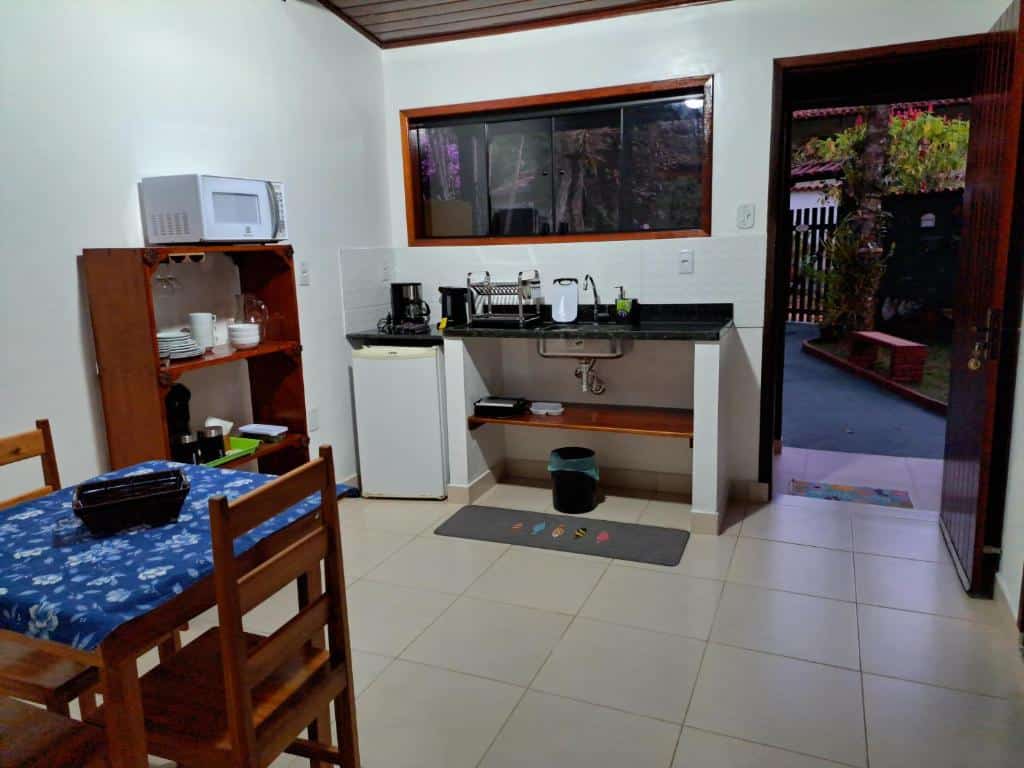Cozinha do airbnb Recanto das Flores Lofts. No lado direito há uma porta aberta, que dá acesso a saída do airbnb, no lado esquerdo há uma mesa com duas cadeiras, um armário encostado na parede e atrás há a pia com um frigobar ao lado.