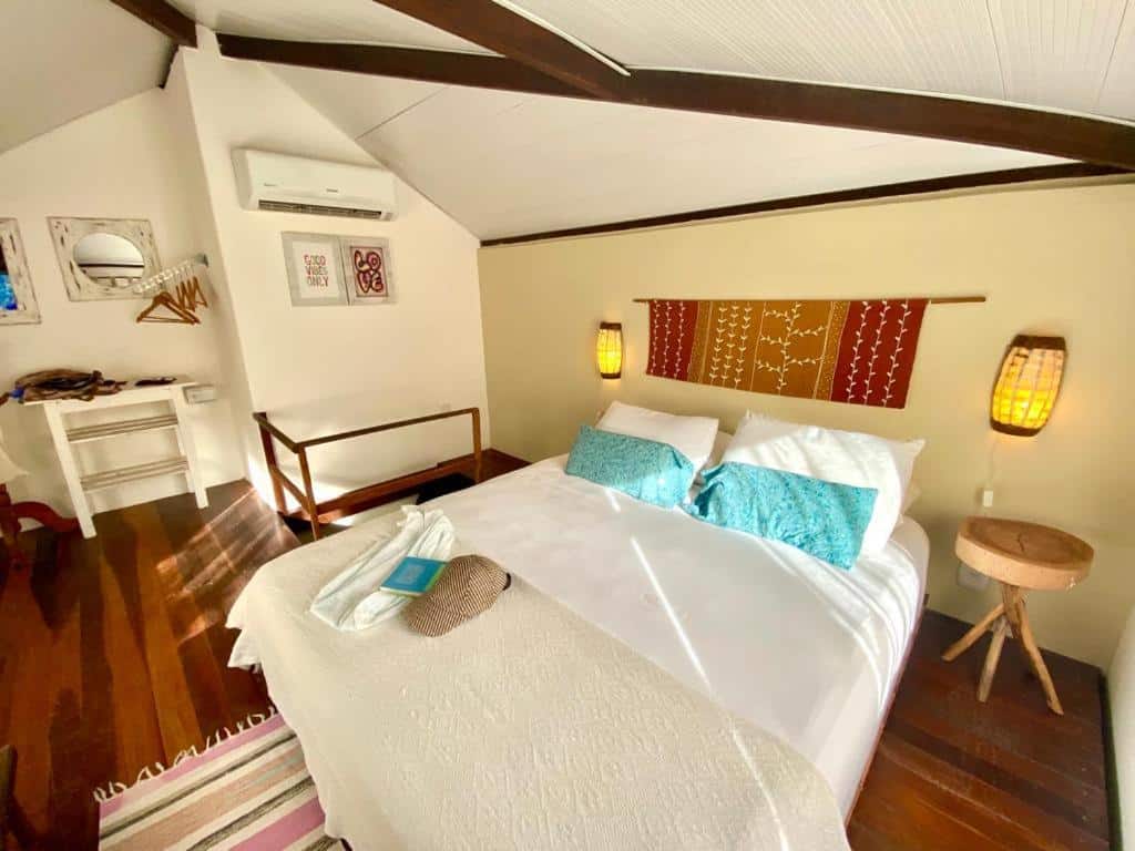 Foto do quarto do Cumelen. Representa o post sobre airbnb em Jericoacoara. Há uma cama de casal e box no centro, possuindo um abajur em seus dois lados. No fundo à esquerda há uma área com mesa, cabides e espelhos.