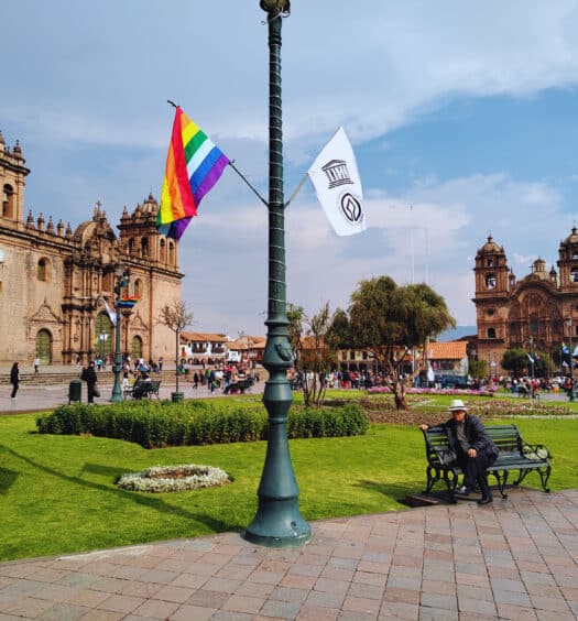 Plaza de las armas de Cusco com um poste de luz bem ao centro, com duas bandeiras de cada lado: a inca e a peruana. Ao fundo é possível ver a Catedral de Cusco à esquerda e a Iglesia de la compañia de Jesus à direita.