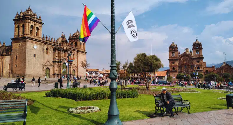 Plaza de las armas de Cusco com um poste de luz bem ao centro, com duas bandeiras de cada lado: a inca e a peruana. Ao fundo é possível ver a Catedral de Cusco à esquerda e a Iglesia de la compañia de Jesus à direita.