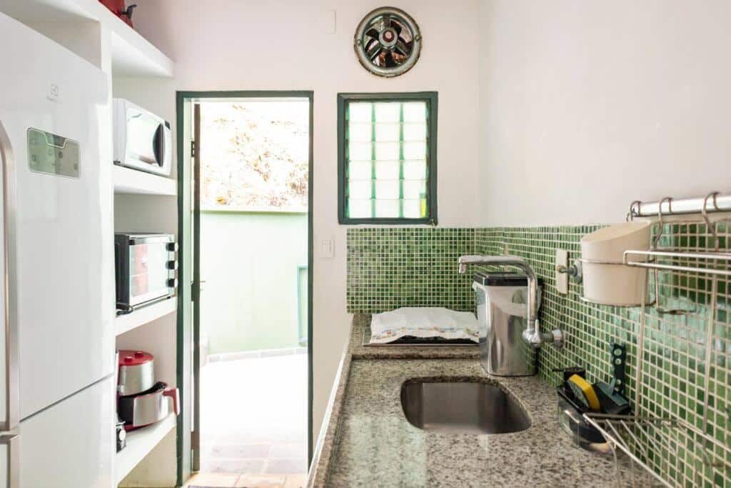 Cozinha do Delba Costa Smeralda. Ao lado direito está a pia cercada por utensílios, e logo atrás um armário com microóndas, forno e air fryer está ao lado de uma geladeira. O corredor entre os móveis termina em uma porta.