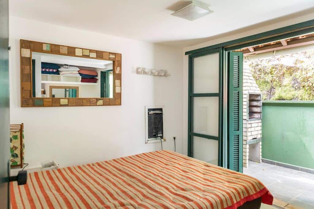 Quarto do Delba Costa Smeralda, um dos airbnb em Barra do Una. Uma cama de casal encara uma porta aberta para a varanda com churrasqueira na parede em frente. Ao lado esquerdo, um espelho reflete a imagem de um armário com travesseiros e cobertores.