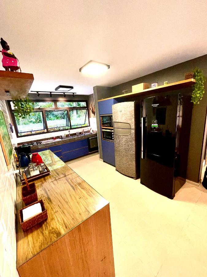 Cozinha da Delícia de casa em Barra do Una a 5 min de Juquehy! Ao lado esquerdo está uma bancada de madeira com comodidades para fazer chá e café, e logo em frente estão duas geladeiras e um apoio com micro-ondas. Ao fundo há uma bancada com pia e fogão, com várias janelas logo acima.