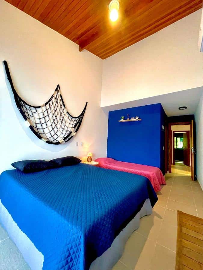 Quarto na Delícia de casa em Barra do Una a 5 min de Juquehy!, um dos airbnb em Barra do Una. Uma cama de casal e uma de solteiro são separadas por uma mesinha com abajur. Na parede atrás da cama está uma rede pendurada.