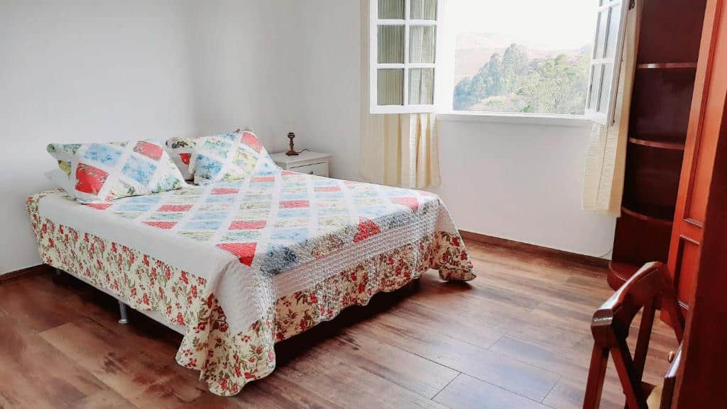 Foto do quarto do Dikasa Alugue Temporada. Ilustra o post sobre airbnb em Ouro Preto. A cama de casal está à esquerda, e na frente dela vemos de relance uma escrivaninha e armário. Do lado direito da cama há uma janela com vista para as montanhas.