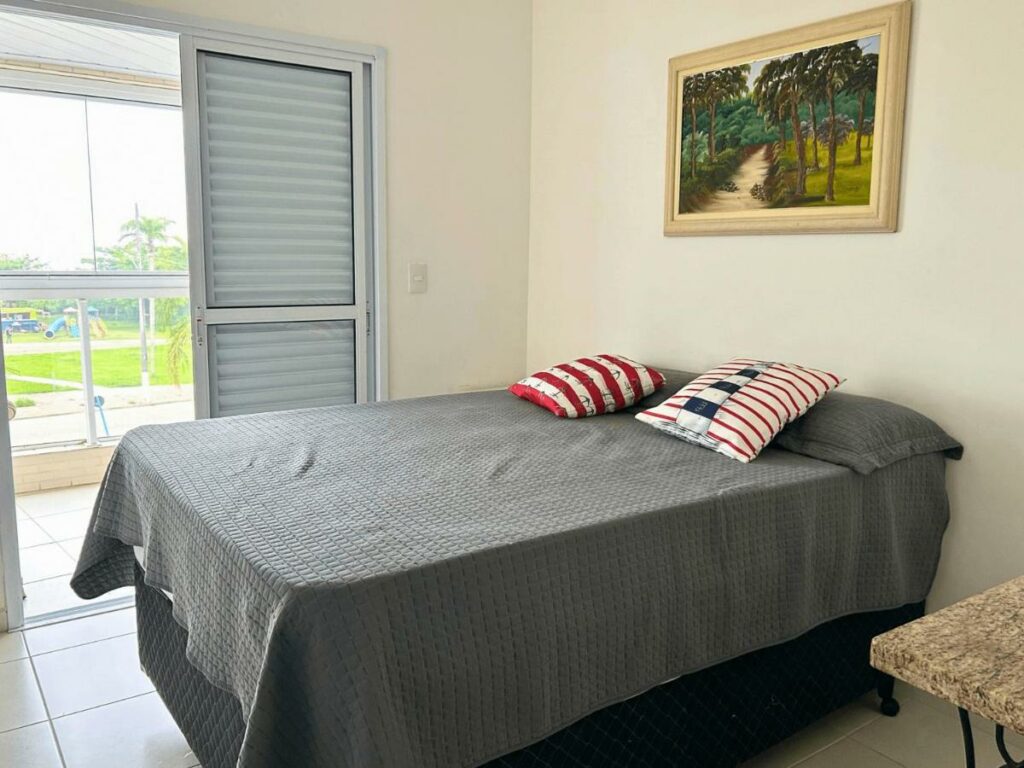 Quarto do Doce Lar de Bertioga Frente ao Mar (Indaiá), um dos airbnb em Bertioga. Uma cama de casal está encostada na parede ao lado direito com um quadrinho logo acima. Na parede ao lado há uma porta com vista para a varanda e um parquinho ao longe.