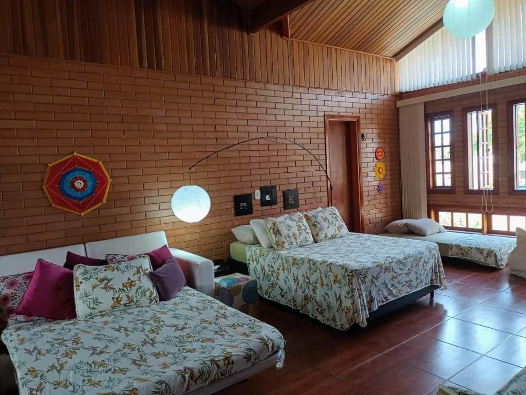 Quarto da EccO Capricornio, um dos airbnb em Caraguatatuba. Um sofá cama com almofadas está aberto ao lado esquerdo da cama de casal, que tem um abajur ao lado. Ao lado direito da cama está uma cama de solteiro ao lado de uma janela. Todas as camas têm travesseiros e jogo de cama.