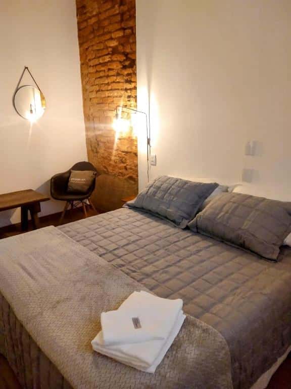 Foto do quarto do Encanto do Pilar. Ilustra o post sobre airbnb em Ouro Preto. A cama é box e de casal, do lado direito dela há uma luminária, um espelho, uma poltrona e um banco de madeira.